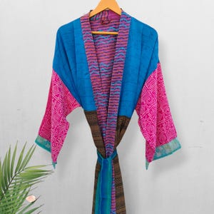 May include: A vibrant, patterned robe in shades of blue, pink, and brown hangs on a wooden hanger. The robe features a geometric design on the sleeves and a contrasting pattern on the lapel and tie. The robe is made of a lightweight fabric.