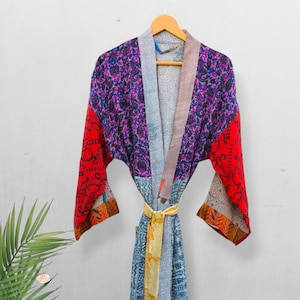 Puede incluir: Una colorida bata de patchwork con un lazo amarillo, colgada de una percha de madera. La bata presenta una mezcla de patrones y colores, incluyendo rojo, morado, azul y gris. Las mangas son rojas con un diseño detallado.