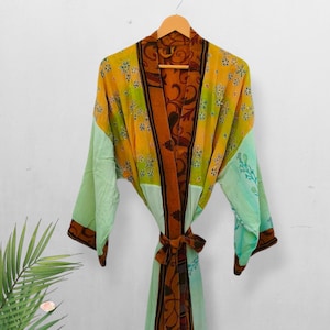 Può includere: Un accappatoio fantasia con cintura annodata, appeso a una gruccia di legno. L'accappatoio presenta tonalità gialle, verdi e marroni con disegni floreali e paisley. Le maniche e la parte inferiore sono verde chiaro.