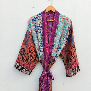 May include: A long-sleeved kimono robe featuring a vibrant patchwork of paisley patterns in shades of magenta, turquoise, orange, and black. The garment includes a matching fabric tie belt at the waist and a wide collar, all hanging on a wooden hanger.