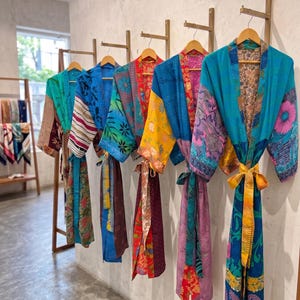 May include: Five long-sleeved robes hanging on wooden wall hooks. Each robe features a unique combination of vibrant, multi-colored fabric panels with various floral, paisley, and geometric patterns, including shades of blue, yellow, pink, and red.