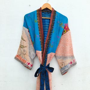 May include: A long-sleeved kimono robe featuring a patchwork design with vibrant blue, peach, and cream panels. The garment includes intricate floral and geometric patterns, a dark brown patterned trim along the front opening, and a solid dark blue tie belt.