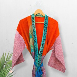 May include: A long-sleeved kimono robe featuring a vibrant orange main body with contrasting light pink sleeves adorned with a white floral pattern. The garment includes a teal trim with a black botanical print and a matching tie belt at the waist.