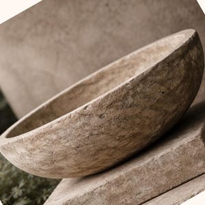 Puede incluir: Un cuenco de piedra ovalado hecho a mano con una superficie rugosa y texturizada. El cuenco es de color marrón claro con vetas marrones más oscuras. El cuenco descansa sobre una superficie de piedra. El tema general es la decoración rústica del hogar.