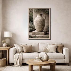 May include: A framed print depicting a large, textured, beige vase with a handle, set on a stone block. The background features greenery and a column. The artwork is displayed in a living room with a neutral colour palette.