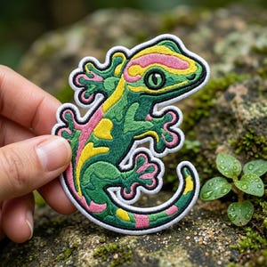 May include: Embroidered patch featuring a colorful gecko design. The gecko has a green body with yellow and pink accents, outlined in white. The patch is held against a natural, mossy background.