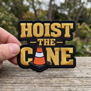 May include: Embroidered patch with the words "HOIST - THE - CONE" in gold thread on a black background. The patch features a traffic cone graphic. The patch is held up by a hand.