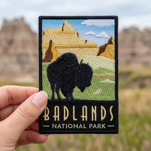 May include: Embroidered patch featuring a black bison in front of a landscape with tan rock formations, green grass, and a blue sky with clouds. The text "BADLANDS NATIONAL PARK" is in gold lettering.