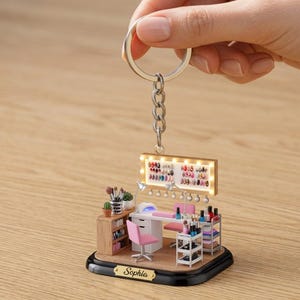 May include: Miniature nail salon keychain with a silver keyring. The scene includes a nail polish display, a manicure table, a pink chair, and a small shelf. The base is black with the name "Sophia" engraved on a gold plaque.