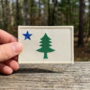 May include: Rectangular patch featuring the flag of Maine. The patch has a cream-colored background with a blue star in the upper left corner and a green pine tree in the center. The patch is bordered with a cream-colored stitching.