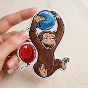 May include: Embroidered patch featuring a cartoon monkey holding a blue balloon overhead and a red balloon below. The monkey is brown with a smiling face and tan hands and feet. The patch has a white border.