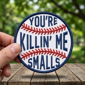 May include: A round baseball-themed patch with the text "YOU'RE KILLIN' ME SMALLS" in navy blue, surrounded by red stitching, on a white background. The patch has a navy blue border. The patch is held in a hand.