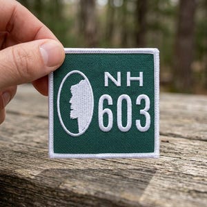 May include: A square, dark green patch with white embroidered text and design. The patch features the letters "NH" above the number "603". A white oval contains a white silhouette of a mountain. The patch is held by a hand.
