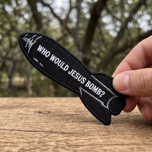 May include: Black embroidered patch in the shape of a rocket. The patch features the text "WHO WOULD JESUS BOMB?" in white lettering. The patch is held against a blurred natural background.