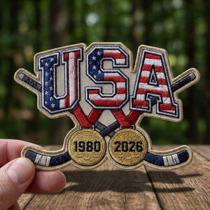 May include: Embroidered patch featuring the word "USA" in red, white, and blue, with a hockey stick on each side. Below the letters are two gold medals with the years "1980" and "2026".