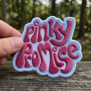 May include: Embroidered patch with the words "Pinky Promise" in a retro font. The text is a vibrant pink color, outlined in a light blue border, and set against a white background. The patch is likely for clothing or accessories.