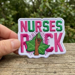 May include: Embroidered patch with the words "NURSES ROCK" in pink and green letters. The patch features a hand with a leaf design and a white border. The patch is on a wooden surface.