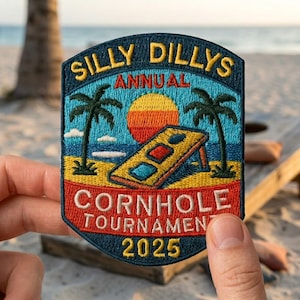 May include: Embroidered patch with the text "SILLY DILLYS ANNUAL CORNHOLE TOURNAMENT 2025". The patch features a beach scene with palm trees, a sunset, and a cornhole board. The patch is blue, red, yellow, and orange.