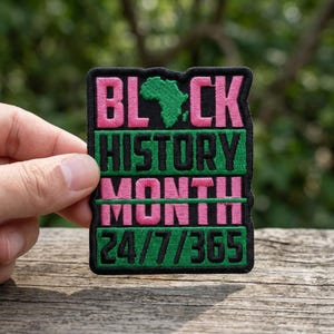 May include: A rectangular patch with the words "BLACK HISTORY MONTH 24/7/365" in pink and green. The word "BLACK" has a green outline of the African continent. The patch has a black border and is on a wooden surface.