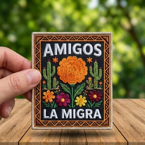 May include: Embroidered patch with the words "AMIGOS LA MIGRA" in white on a black background. The patch features a large orange flower, cacti, and colorful flowers. The patch is held by a hand.