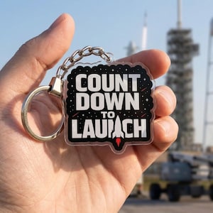 May include: A clear acrylic keychain with the words "COUNT DOWN TO LAUNCH" in white lettering on a black background. A red rocket graphic is at the bottom. The keychain has a silver keyring and chain.