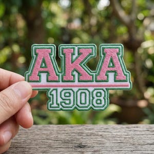 May include: Embroidered patch with the letters "AKA" in pink, outlined in green, above the year "1908" in green. The patch is held against a blurred green background and a wooden surface.