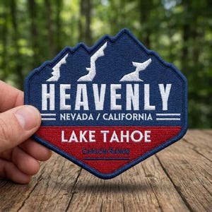 May include: Embroidered patch with a blue mountain design and the word "HEAVENLY" in white. Below, it reads "NEVADA / CALIFORNIA" and "LAKE TAHOE" in white and red, respectively. The patch is held against a wooden surface.