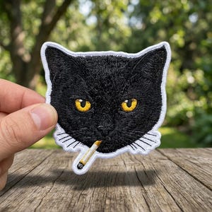 May include: Embroidered patch featuring a black cat face with yellow eyes and a cigarette in its mouth. The patch has a white border and is held against a blurred green background and a wooden surface.