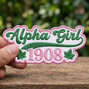 May include: Embroidered patch with the words "Alpha Girl 1908" in green and pink. The patch has a white background with a pink border and green leaf accents. The patch is held up against a blurred green background.