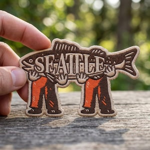 May include: Embroidered patch featuring a fish design with the word "SEATTLE" in the center. The fish is brown with orange pants and brown shoes. The patch is held up by two figures. The patch is on a wooden surface.
