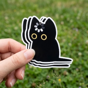 May include: Black cat sticker with a white outline, featuring yellow eyes and a loading symbol on its head. The sticker is held against a blurred green background, suggesting an outdoor setting.