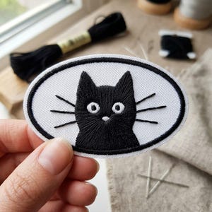 May include: An oval-shaped, embroidered patch featuring a black cat face with white eyes and whiskers. The cat is centered on a white background with a black border. The patch is held in a hand.