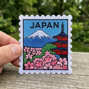 May include: Embroidered patch featuring a Japanese landscape design. The patch depicts Mount Fuji, a pagoda, and cherry blossoms, with the word "JAPAN" at the top. The patch has a white border and a blue sky background.