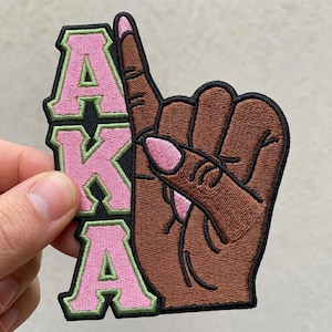 May include: Embroidered patch featuring a brown hand with a pinky nail and the letters "AKA" in pink with green trim. The patch is on a black background.