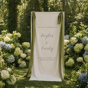 May include: A cream wedding sign with green ribbons. Text reads "WELCOME TO THE WEDDING OF Jaylas & Emily SATURDAY, OCTOBER. 2, 2026 MITTENWALD, GERMANY". The sign is set in a garden with flowers and trees.