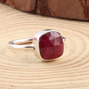 Pu&ograve; includere: Un anello d'argento con una gemma rosso scuro, tagliata a forma quadrata. L'anello &egrave; posto su una superficie di legno chiaro con un bordo marrone scuro. La gemma &egrave; il punto focale, mostrando il suo colore e taglio.