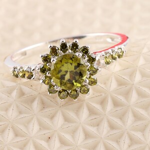 May include: A silver ring featuring a central, round, olive-green gemstone surrounded by smaller, matching stones in a floral design. Additional small green stones are set along the band, creating a sparkling, elegant piece of jewelry.