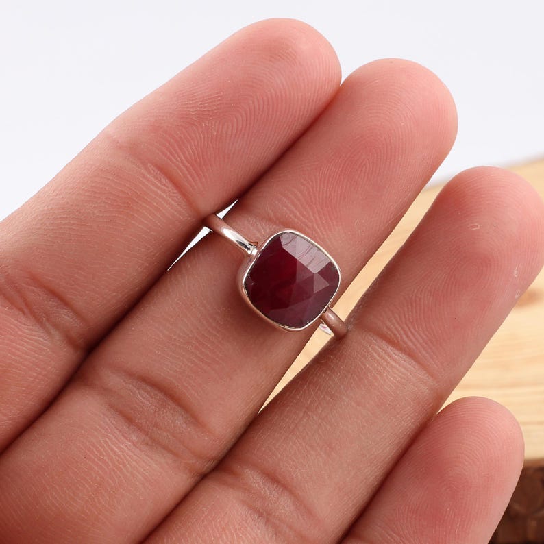 Pu&ograve; includere: Un anello d'argento con una gemma rosso scuro a taglio cuscino. La gemma &egrave; incastonata in una fascia semplice ed elegante. L'anello &egrave; esposto su una superficie chiara.