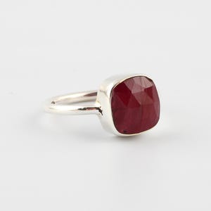 Pu&ograve; includere: Un anello d'argento con una grande gemma di rubino a taglio quadrato. Il rubino &egrave; di un colore rosso intenso e ricco, e l'incastonatura &egrave; una semplice fascia d'argento lucida. L'anello &egrave; esposto su sfondo bianco.