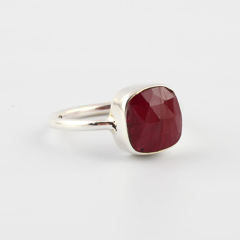 Pu&ograve; includere: Un anello d'argento con una grande gemma di rubino a taglio quadrato. Il rubino &egrave; di un colore rosso intenso e ricco, e l'incastonatura &egrave; una semplice fascia d'argento lucida. L'anello &egrave; esposto su sfondo bianco.