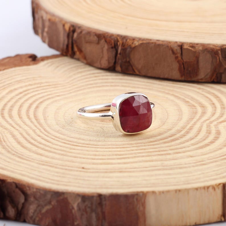 Pu&ograve; includere: Un anello d'argento con una gemma rosso scuro, tagliata a forma quadrata. L'anello &egrave; posto su una superficie di legno chiaro con un bordo marrone scuro. La gemma &egrave; il punto focale, mostrando il suo colore e taglio.