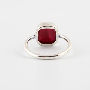 Pu&ograve; includere: Un anello d'argento con una gemma rosso scuro quadrata. La gemma &egrave; incastonata in una montatura a castone e la fascia &egrave; liscia e lucida. L'anello &egrave; mostrato su sfondo bianco. Questo &egrave; un gioiello.