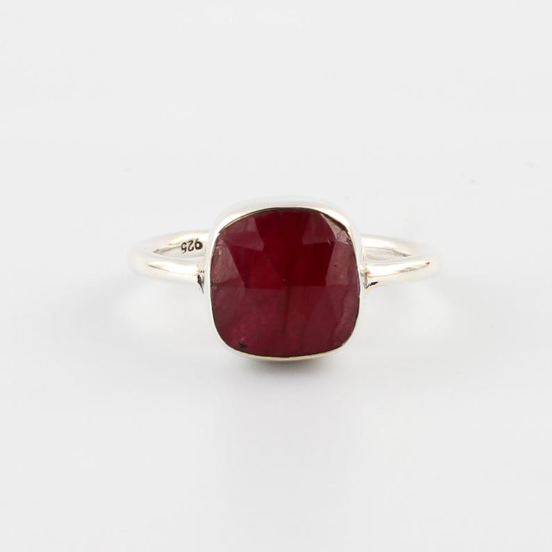 Pu&ograve; includere: Un anello d'argento con una grande pietra preziosa rosso scuro a taglio quadrato. La gemma sfaccettata cattura la luce. La fascia dell'anello &egrave; sottile e lucida, con un piccolo segno vicino alla gemma. L'anello &egrave; posto su uno sfondo bianco.