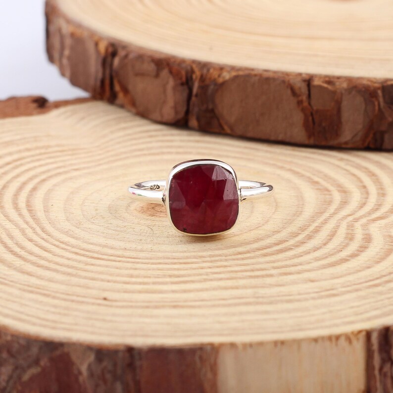 Pu&ograve; includere: Un anello d'argento con una grande pietra preziosa rosso scuro a taglio quadrato. L'anello &egrave; appoggiato su una superficie di legno con un motivo a venature naturali. La pietra preziosa &egrave; il punto focale, catturando la luce e aggiungendo eleganza.