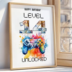 May include: A framed print with the text "HAPPY BIRTHDAY LEVEL 14 UNLOCKED." The number 14 is filled with images of money. A watercolor-style game controller is below the text, with blue, yellow, and rainbow colors.