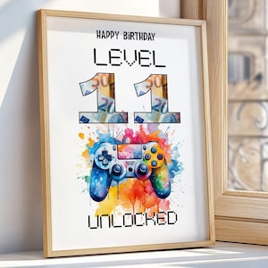 Puede incluir: Impresión enmarcada con el texto "Happy Birthday LEVEL 11 UNLOCKED". El número 11 está diseñado con moneda. Una imagen de estilo acuarela de un mando de juego está debajo del texto, con salpicaduras de pintura coloridas.