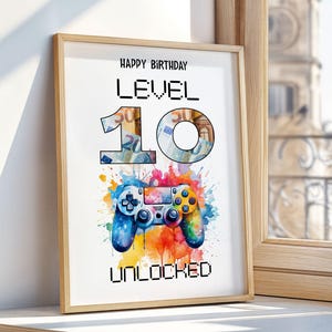 Könnte beinhalten: Gerahmter Druck mit der Aufschrift "Happy Birthday Level 10 Unlocked". Die Zahl 10 ist mit Bildern von Geld gefüllt. Unten befindet sich ein farbenfrohes Gamepad im Aquarellstil mit einem lebendigen Farbspritzer.