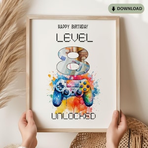 Könnte beinhalten: Gerahmter Druck mit dem Text "Happy Birthday Level 8 Unlocked". Die Zahl 8 ist mit einem Gelddesign gefüllt, und ein farbenfroher Gamecontroller im Aquarellstil befindet sich unten. Der Druck wird von Händen gehalten.