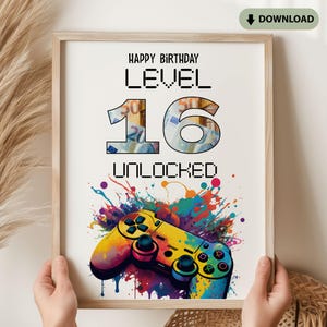 Op de afbeelding: Een ingelijste print met de tekst "HAPPY BIRTHDAY LEVEL 16 UNLOCKED". De cijfers zijn gevuld met een gelddesign. Onderaan staat een kleurrijke gamecontroller met verfspattereffecten. De print wordt door handen vastgehouden.