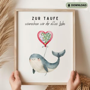 May include: A framed print featuring a watercolor illustration of a blue whale holding a red heart-shaped balloon containing a 100 Euro bill. The text reads ZUR TAUFE wünschen wir dir alles Liebe. The frame is light wood with a white background.